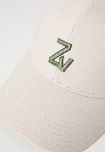 Gorra de béisbol beige hecha de tela texturizada, con un logo bordado en verde y detalles de costura en el ala y la corona.