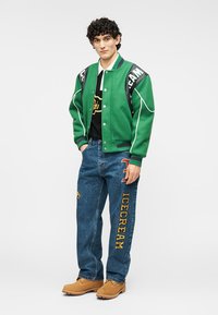 Πράσινο varsity μπουφάν με λευκή περίγραμμα, μαύρες λεπτομέρειες, μαύρο κοντομάνικο μπλουζάκι, μπλε τζιν παντελόνι με κέντημα "ICECREAM" και λογότυπο, καφέ παπούτσια.