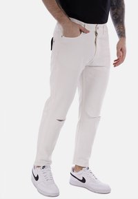 Jeans in denim bianco con vestibilità slim, caratterizzati da patta con zip e tasche frontali, dettagli strappati sulle ginocchia, abbinati a sneakers bianche.