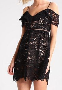 Vestido negro de encaje con detalles de volantes sin mangas, patrón floral y cintura ajustada. Tela transparente con dobladillo en forma de concha.