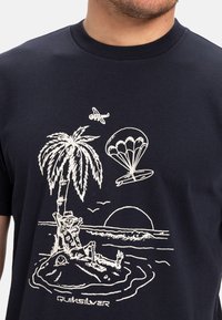 Navy blue t-shirt featuring white graphic design of a palm tree, a skeleton in a chair, a parachute, and a sunset over water.
