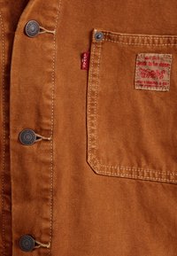 Brązowa kurtka Levi's z metalowymi guzikami, przednią kieszenią, czerwoną metką Levi's oraz naszywką z napisem "gdy jest do zrobienia praca".