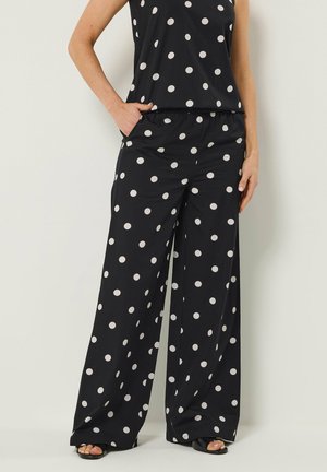 Schwarze weit geschnittene Hose mit weißen Punkten, elastischem Bund, Seitentaschen, getragen mit passendem ärmellosem Oberteil und schwarzen Sandalen mit offenen Zehen.