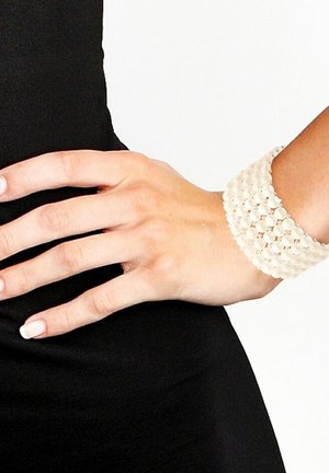 Bracelet large et blanc en perles entrelacées avec une finition texturée, porté sur une main. Comprend un fermoir avec un accent métallique lisse.