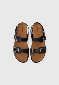 Zwarte leren sandalen met twee verstelbare gespen, een zachte bruine suède voetbed en een vlakke rubberen zool. De bovenaanzicht laat alle ontwerpelementen duidelijk zien.