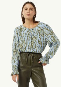Bluse mit floralem Muster in Blau- und Grüntönen, ausgestattet mit einem runden Ausschnitt, Raffung und weichem Stoff. Kombiniert mit dunkelgrünen Hosen.