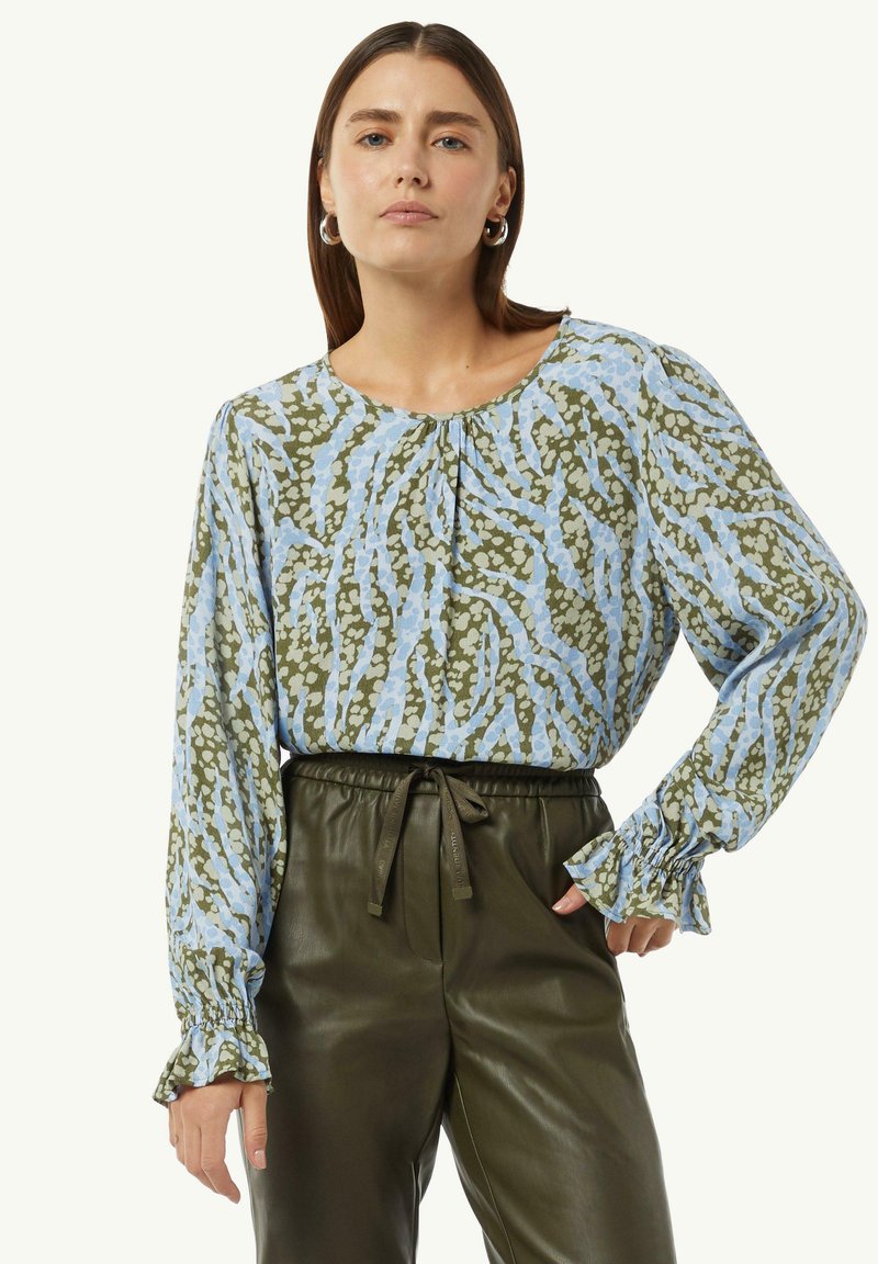 Bluse mit floralem Muster in Blau- und Grüntönen, ausgestattet mit einem runden Ausschnitt, Raffung und weichem Stoff. Kombiniert mit dunkelgrünen Hosen.