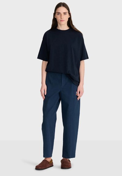 Femme aux longs cheveux bruns portant un t-shirt noir ample, un pantalon bleu marine, des chaussettes rayées et des chaussures marron sans lacets, debout devant un fond uni.
