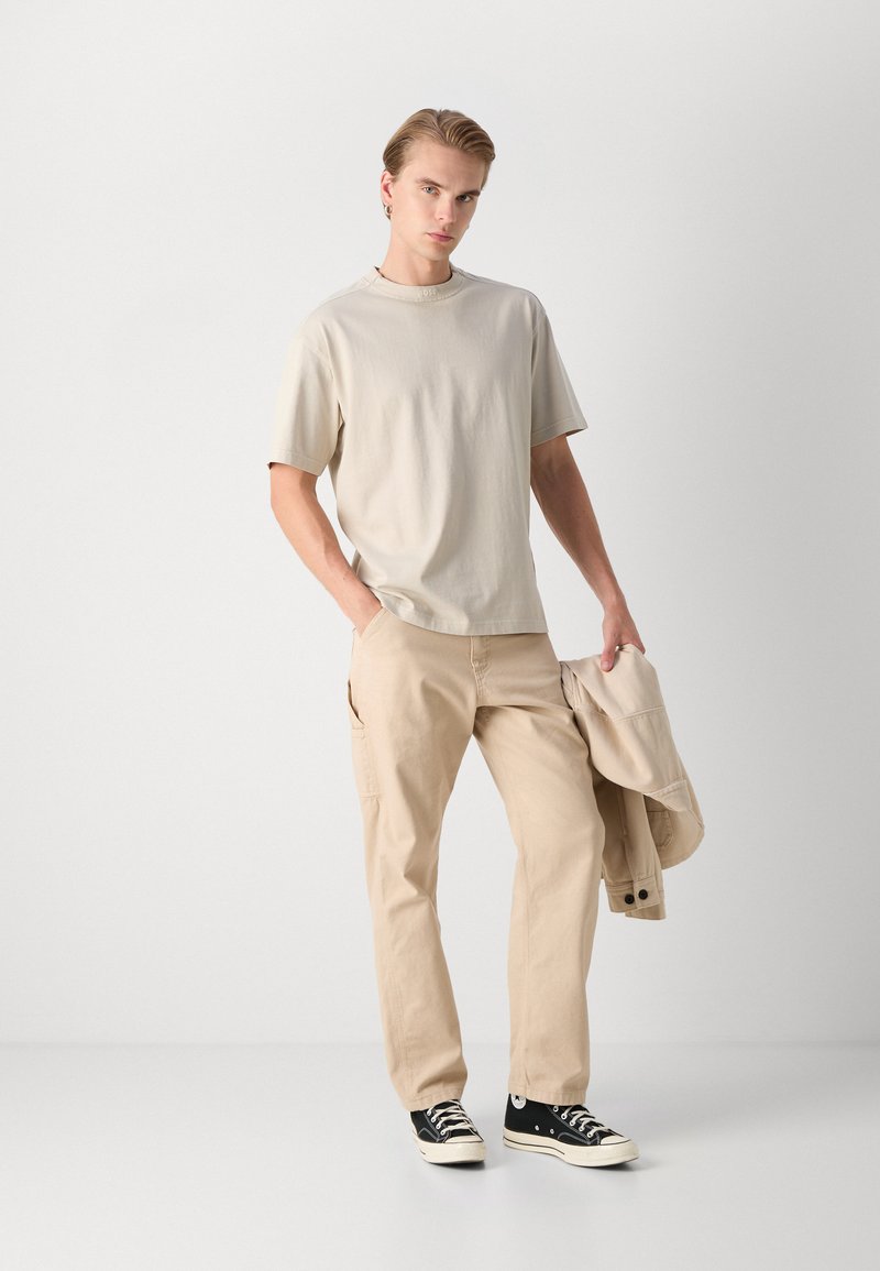 Beige katoenen t-shirt, ontspannen pasvorm; beige cargo broek met zijzakken; zwarte canvas sneakers. Model houdt een bijpassende jas vast.
