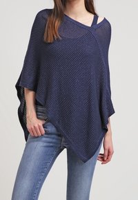 Marinblå stickad poncho med öppen väv, som har en asymmetrisk skärning och vida ärmar. Bärs över en ljus topp och kombineras med smala jeans.