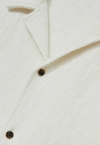 Biała teksturowana kurtka z wzorzystym materiałem, z kołnierzem typu lapel i okrągłym zapięciem na guzik. Tekstura dodaje głębi projektowi.