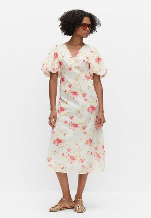 Kvinde med krøllet hår iført orange solbriller og en hvid midikjole med rødt blomsterprint og puffede korte ærmer, stående med hænderne langs siderne.