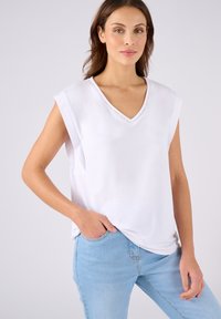 Top bianco senza maniche con scollatura a V, presenta una leggera cucitura lungo il collo. Abbinato a jeans in denim azzurro chiaro. Tessuto morbido.