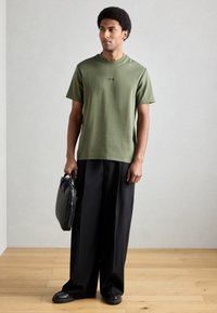 Olivzaļš kokvilnas T-krekls ar īsām piedurknēm, apaļu izgriezumu un smalku izšuvumu, kopā ar melnām plašām biksēm un melnu somu.