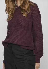 Burgundy stickad tröja med ett strukturerat mönster, långa ärmar och ribbade muddar, tillsammans med svarta byxor. Har en avslappnad passform och rund halsringning.