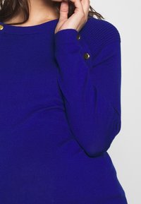 Kungablå stickad tröja med ribbade axlar och guldknappdetaljer, med långa ärmar och en figurnära silhuett.