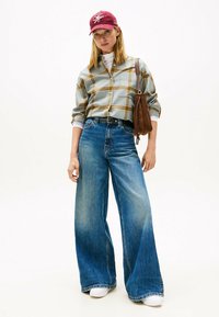 Jeans en denim bleu à jambes larges avec une finition délavée, associés à une chemise à carreaux verte et marron. Une casquette bordeaux et un sac à bandoulière marron complètent le look.