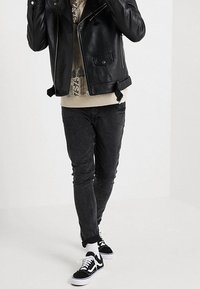 Giacca da moto in pelle nera con zip frontale, abbinata a una camicia beige e jeans in denim scuri. Sneakers nere con suole bianche completano il look.