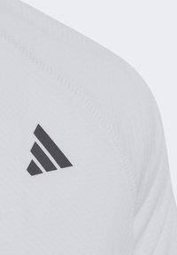 Camiseta deportiva blanca con tejido texturizado y un logo triangular negro en el hombro. Presenta un diseño limpio y detalles de costura visibles.