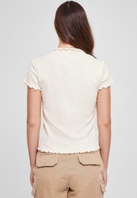 Kortærmet top i bomuld i off-white med bølgede kanter ved hals og lukning. Glat tekstur, tætsiddende design, parret med beige cargo bukser.