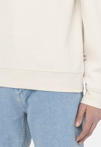 Sudadera de color beige claro con puños y bajo acanalados, combinada con unos jeans de mezclilla azul claro. Pequeña etiqueta de logo en el lateral de la sudadera.