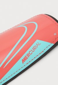 Red shin guard featuring a smooth finish, blue accents, and a branded logo. It has a contoured shape with a black padded edge.