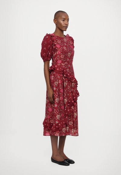 Vestido floral rojo con mangas abullonadas, dobladillo con volantes y patrones intrincados de flores rosas en una tela ligera. Bailarinas negras completan el look.