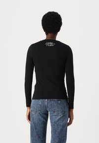 En person med kort lockigt hår bär en svart långärmad topp med vit text vid halsringningen och blå denimjeans mot en enfärgad vit bakgrund.