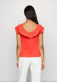 Röd blus med en stor hålmönstrad volang runt halsringningen och en rundad rygg, matchad med vita byxor. Slät textur, åtsittande design.