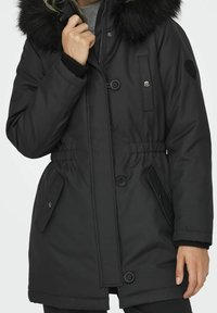 Parka noire isolante avec capuche bordée de fourrure, dotée d'une taille cintrée, de fermetures à boutons et de poches latérales. Tissu doux et lisse.