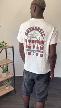 Hvid bomulds t-shirt med rød og grøn print på ryggen, der viser "AUTHENTIC LEVI'S" og et solstråledesign; parret med sorte denim shorts.