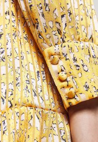 Manche en tissu jaune à motifs avec boutons recouverts de tissu et poignet froncé sur un bras à la peau claire.