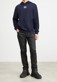 Hombre con sudadera azul marino de Hugo, pantalones negros ajustados y botas de senderismo negras con cordones reflectantes, de pie con una mano en el bolsillo.