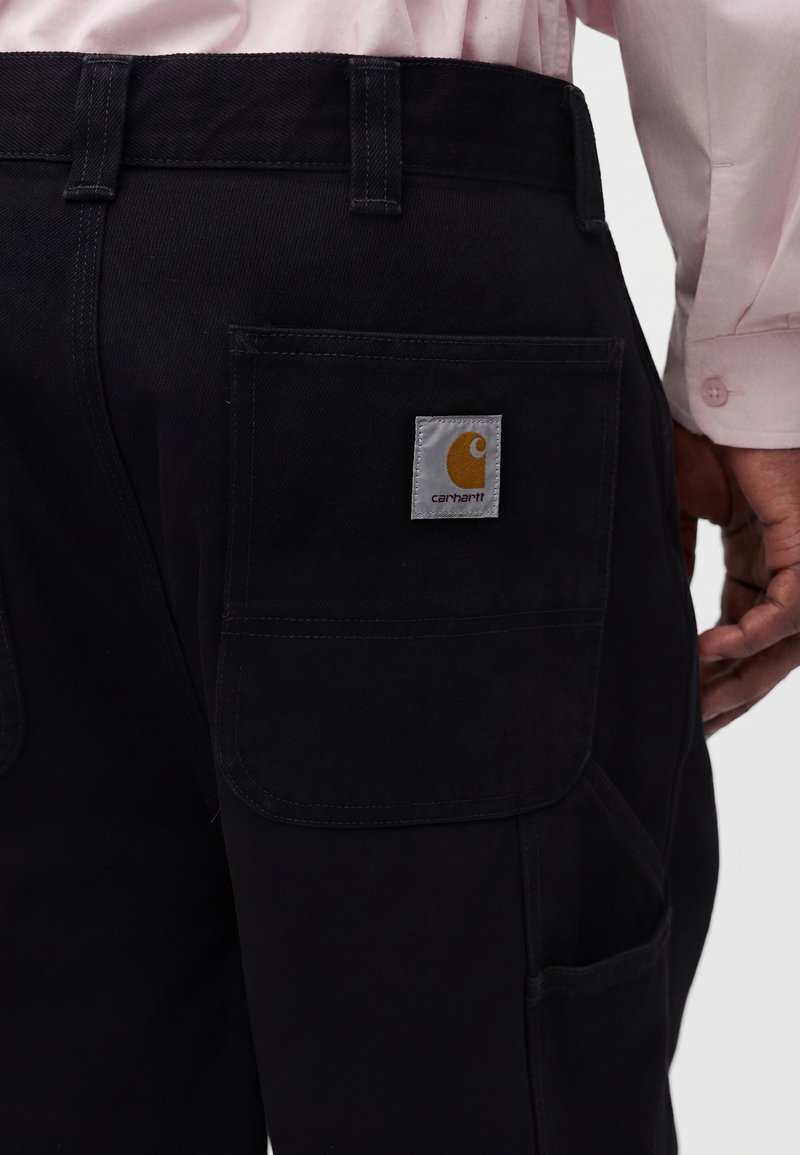 Person trägt schwarze Carhartt-Hose mit einer Gesäßtasche, auf der das Markenlogo zu sehen ist, und ein teilweise sichtbares pinkes Langarmshirt.