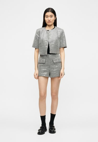 Femme debout portant une veste à manches courtes en pied-de-poule noir et blanc et un short taille haute assorti, des chaussettes noires et des mocassins noirs.