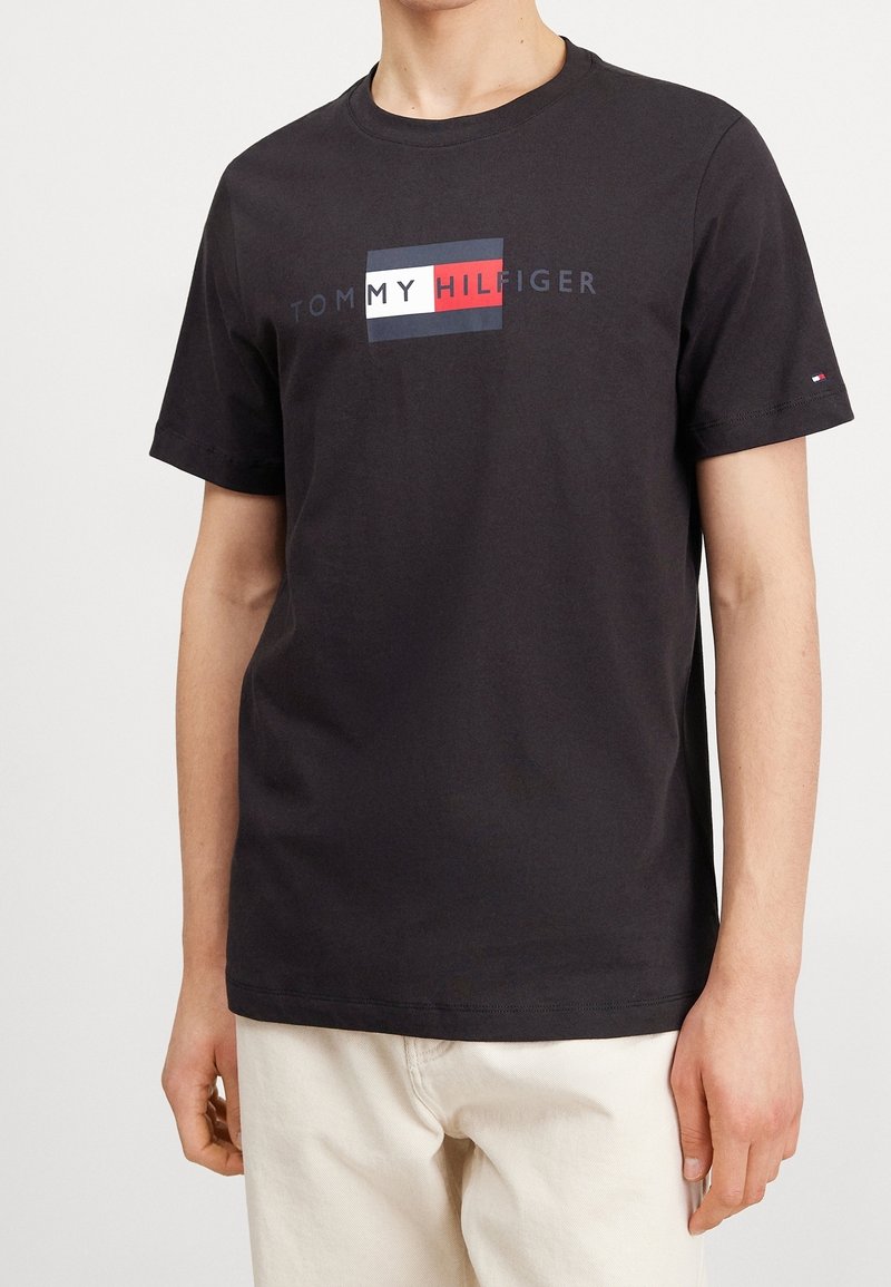 Person wearing a black Tommy Hilfiger t-shirt with logo text and flag design, paired with light beige pants, standing against a plain background.