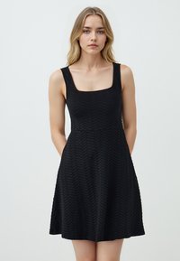 Abito nero senza maniche con scollo quadrato, caratterizzato da un motivo a spina di pesce testurizzato. Silhouette a campana con tasche laterali.