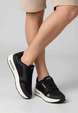 Beine, die beige Shorts und schwarze Giesswein Sneakers mit weißen Sohlen tragen, stehen mit einem Fuß über den anderen auf grauem Hintergrund.