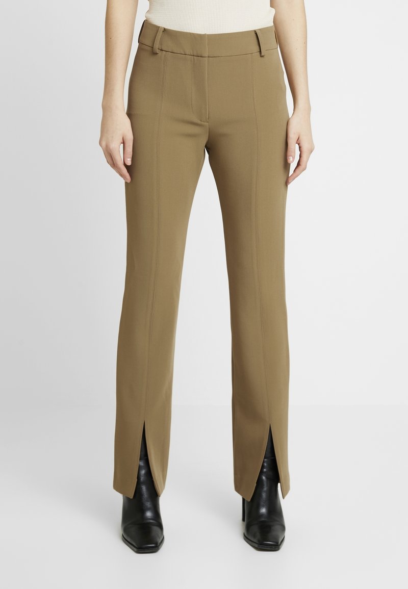 Pantalons marron ajustés avec une taille cintrée, des coutures verticales et des fentes sur les côtés, associés à des bottes à talons noires. Texture de tissu lisse.