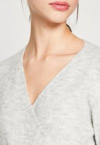 Ljusgrå, strukturerad stickad tröja med V-ringning. Närbildsdetaljer inkluderar mjukt tyg och en avslappnad passform runt halsen och axlarna.