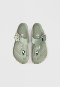 Ανοιχτό πράσινα σανδάλια με ιμάντα που κλείνει με αγκράφα, υφασμάτινη επιφάνεια και μαύρη βάση δακτύλου. Περιλαμβάνουν ένα διακριτικό διακοσμητικό κουμπί στο δάχτυλο.