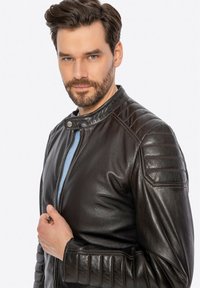 Giacca da motociclista in pelle marrone con dettagli trapuntati sulle spalle, colletto rotondo e zip frontale, indossata da un modello maschile su uno sfondo neutro.