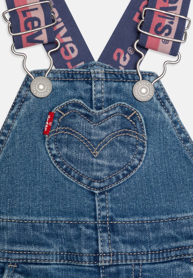 Denim-Trägerhose mit einer herzförmigen Gesäßtasche, dunkelblauen verstellbaren Trägern mit einem Logo und silbernen Hardware-Details.