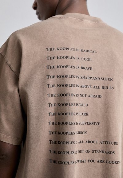 Camiseta de algodón marrón de corte oversize con frases en texto negro impresas verticalmente en la parte posterior. Mangas cortas y un escote redondo clásico.