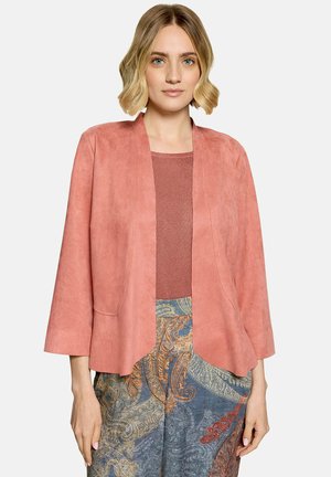 Femme aux cheveux blonds courts porte une veste corail ouverte devant, un haut couleur rouille et un pantalon à motifs cachemire multicolore.