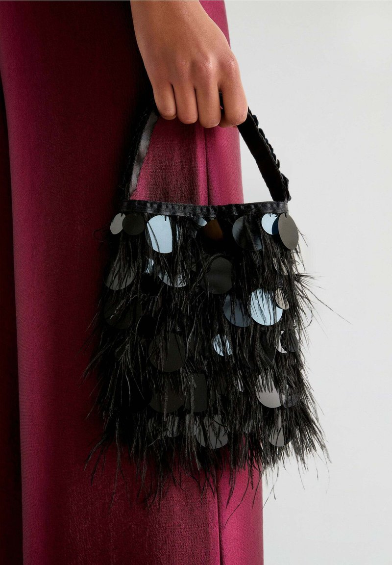Bolso de mano con flecos negros y acentos circulares reflectantes, con una superficie texturizada y un asa trenzada. Colocado sobre un fondo burdeos oscuro.