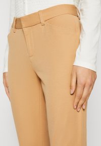 Beige op maat gemaakte broeken van gestructureerde stof, met zijzakken en een gladde tailleband. Hand op het bovenbeen geplaatst.