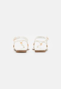 Sandales plates blanches avec des brides tressées, des accents dorés et une semelle beige. Elles possèdent une boucle de cheville ajustable pour un meilleur ajustement.