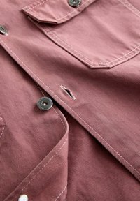 Close-up of unbuttoned pink denim jacket showing metal snap buttons, buttonhole stitching, and front pocket with white thread seams.