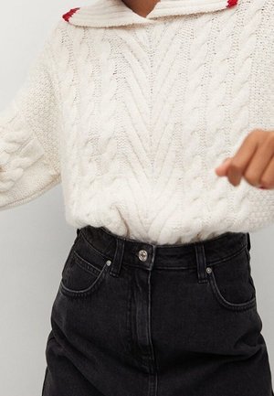 Pull en maille torsadée crème avec un col bordé de rouge, associé à un jean noir taille haute. Texture prononcée et style ajusté.